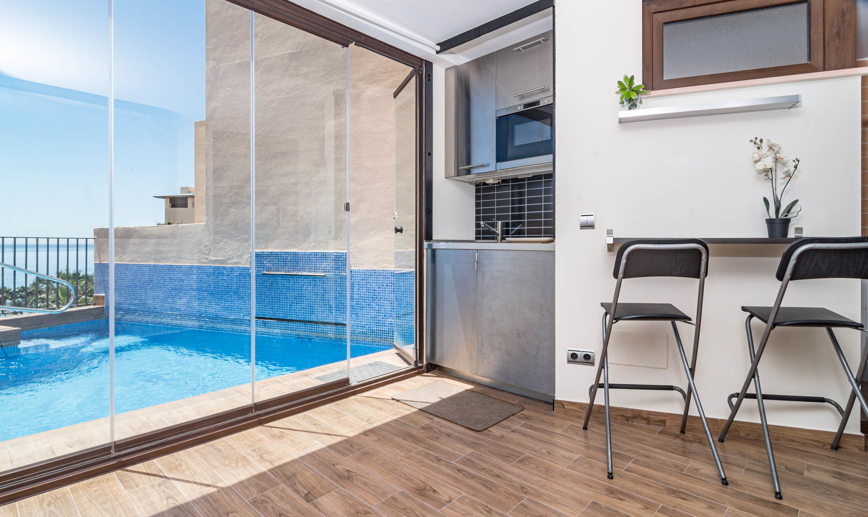 A balcony with a glass door leading to a swimming pool, a small kitchenette, and two chairs.