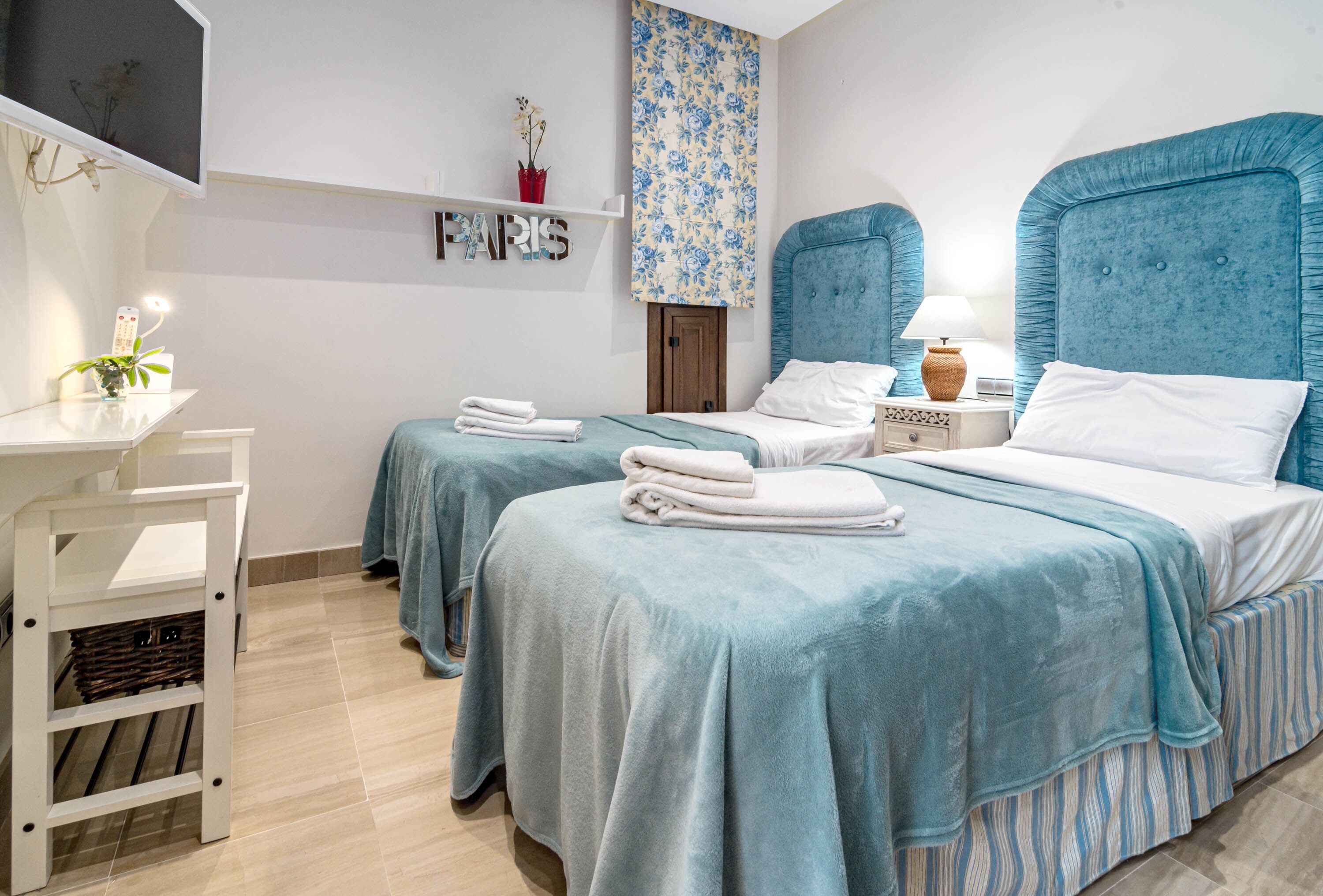 A hotel room with two beds, a small desk, a shelf with a 'PARIS' sign, and a floral-patterned wall hanging.
