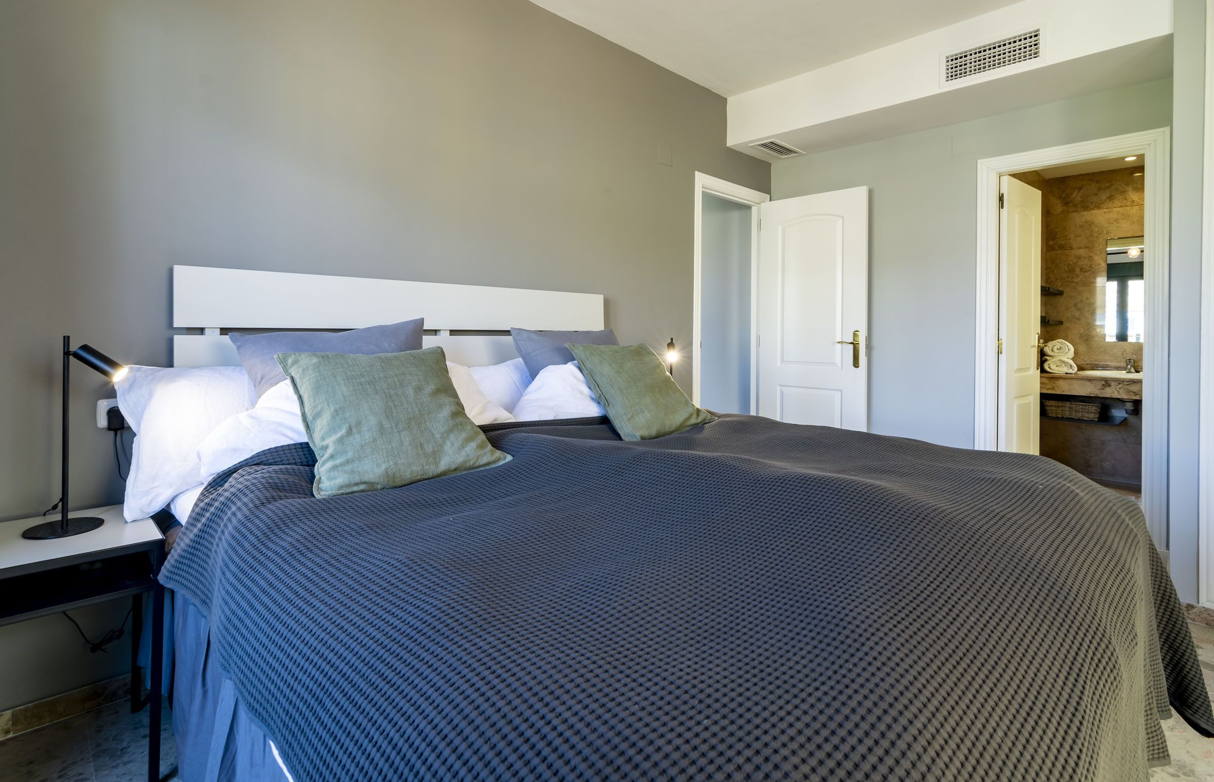 A neatly made bed with a headboard, a bedside table with a lamp, and a door leading to a bathroom.
