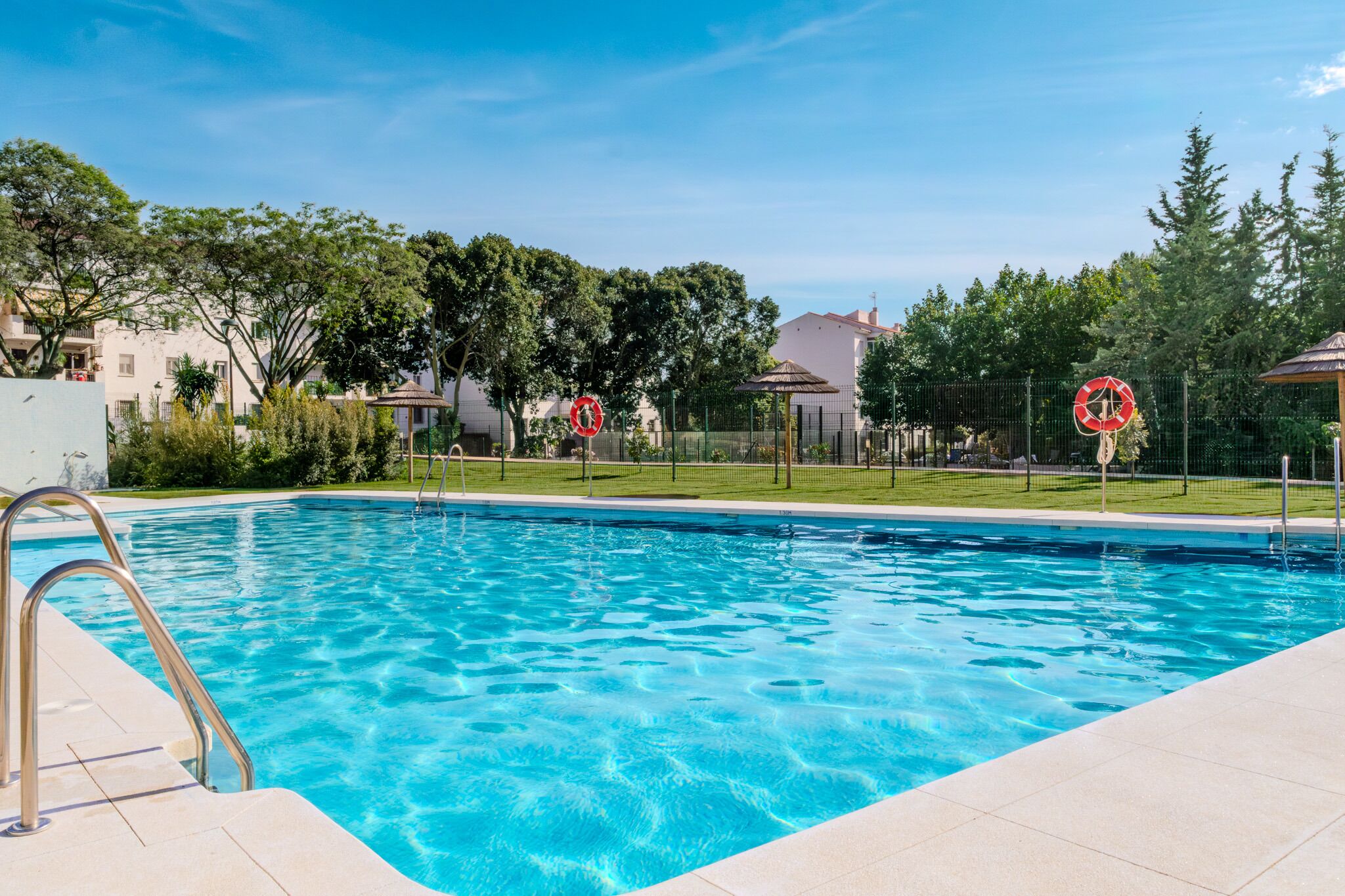 A swimming pool with a metal ladder, surrounded by a fence, trees, and buildings in the background.