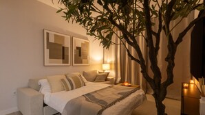 A modern bedroom with a bed, bedside tables, a lamp, a tree, and abstract wall art. - Ae22b - New Build w/ Pool, gym & Sunny Balcony (Estepona)