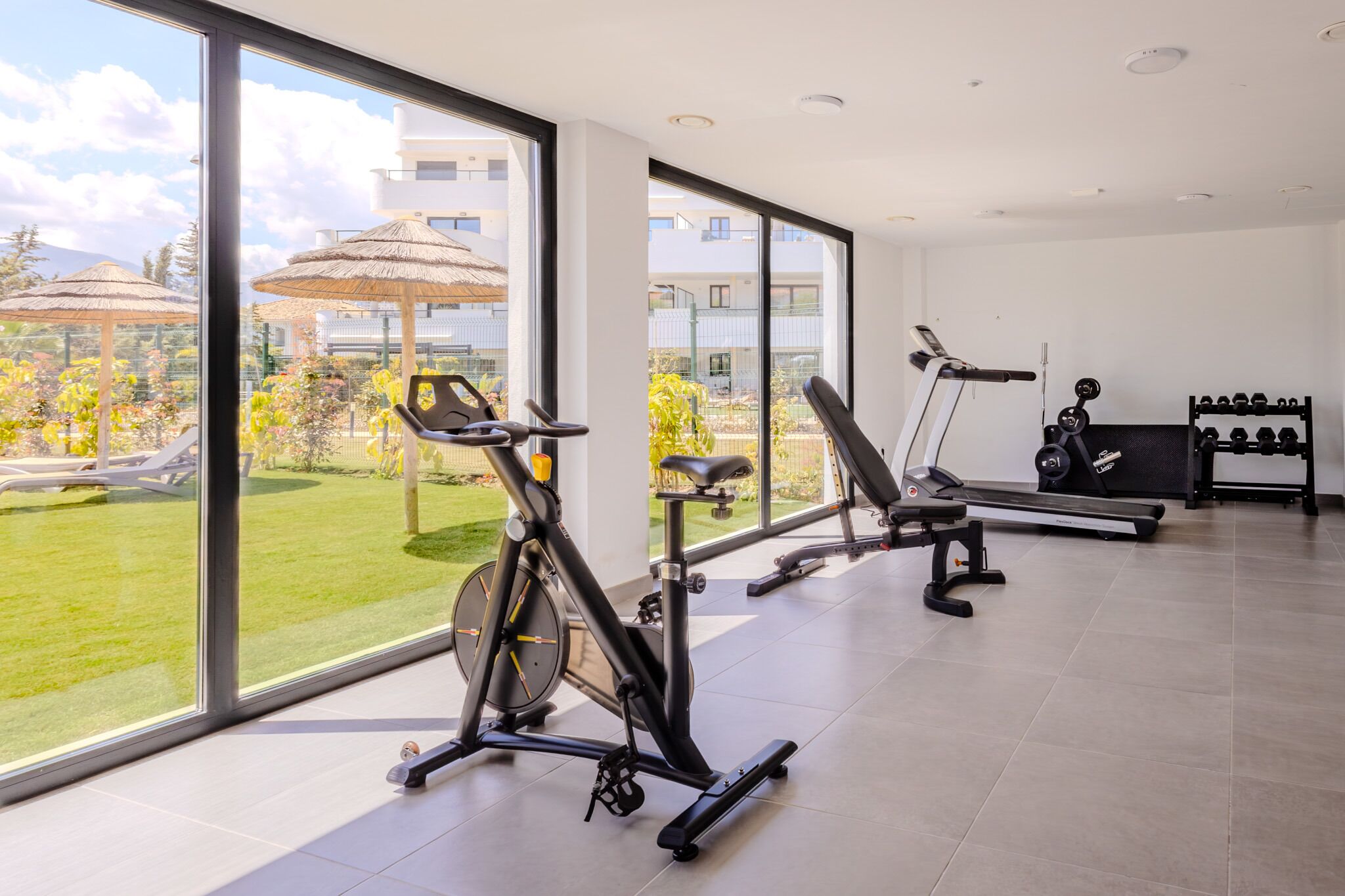 Une salle de sport équipée de matériel d’exercice, dotée de grandes fenêtres et offrant une vue sur un jardin.