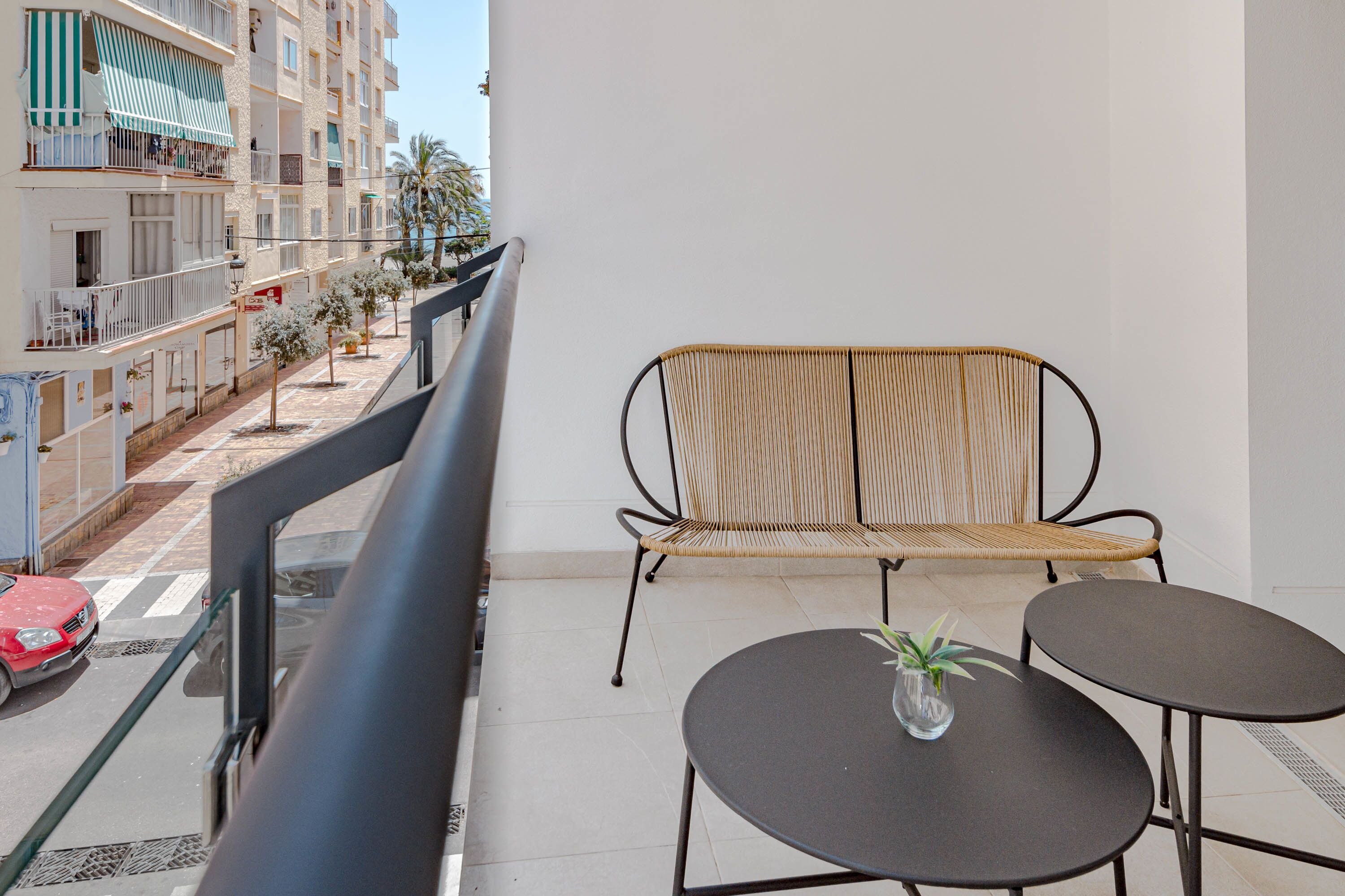 A balcony with a wicker chair and two small tables, a red car parked on the street, and a building with balconies and palm trees in the background.
