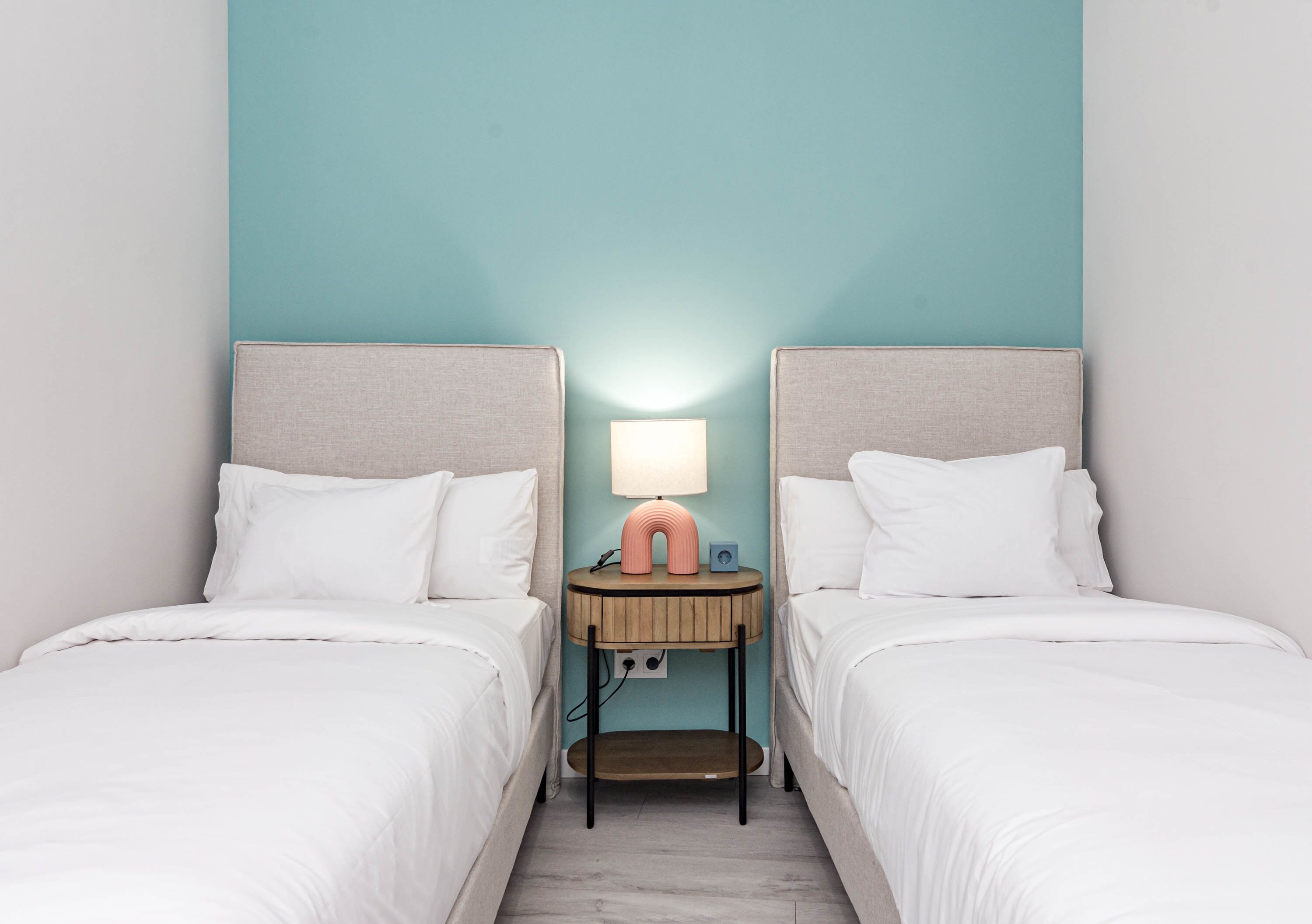 A room with two beds, a nightstand with a lamp, and a wall-mounted outlet.