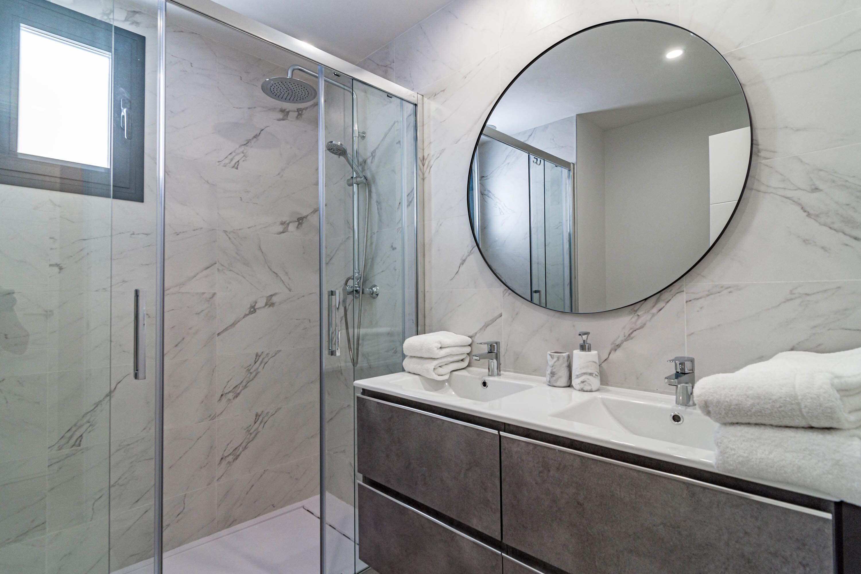 A modern bathroom with a glass-enclosed shower, a round mirror, and a vanity with a sink and towel stack.