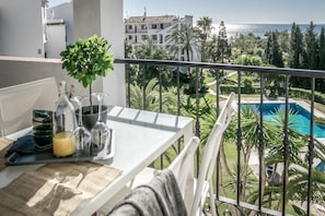 A balcony with a table set for two, a small potted plant, and a view of a swimming pool and surrounding greenery. - RDM - Stylish Holiday Apartment With Ocean Views (Mijas)