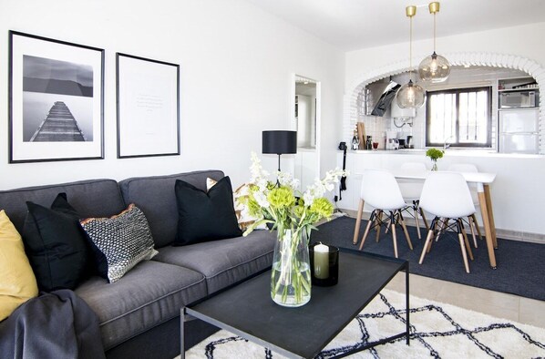 Uma sala de estar moderna com um sofá cinzento, uma mesa de centro e um vaso de flores. Há uma área de jantar com cadeiras brancas e uma mesa. Cozinha visível ao fundo, com armários brancos e eletrodomésticos de aço inoxidável.