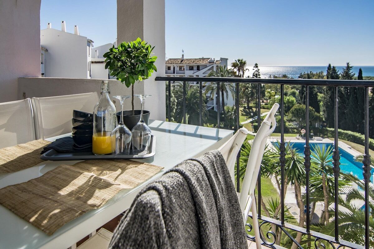 Un balcon avec une table préparée pour le petit-déjeuner, une plante en pot et une vue sur une piscine et l’océan.