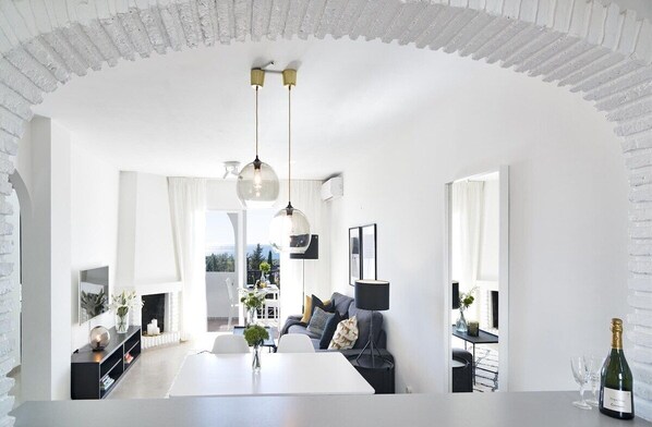 A modern living room with a white fireplace, a dining area, and a view of the outdoors. - RDM - Stylish Holiday Apartment With Ocean Views (Mijas)