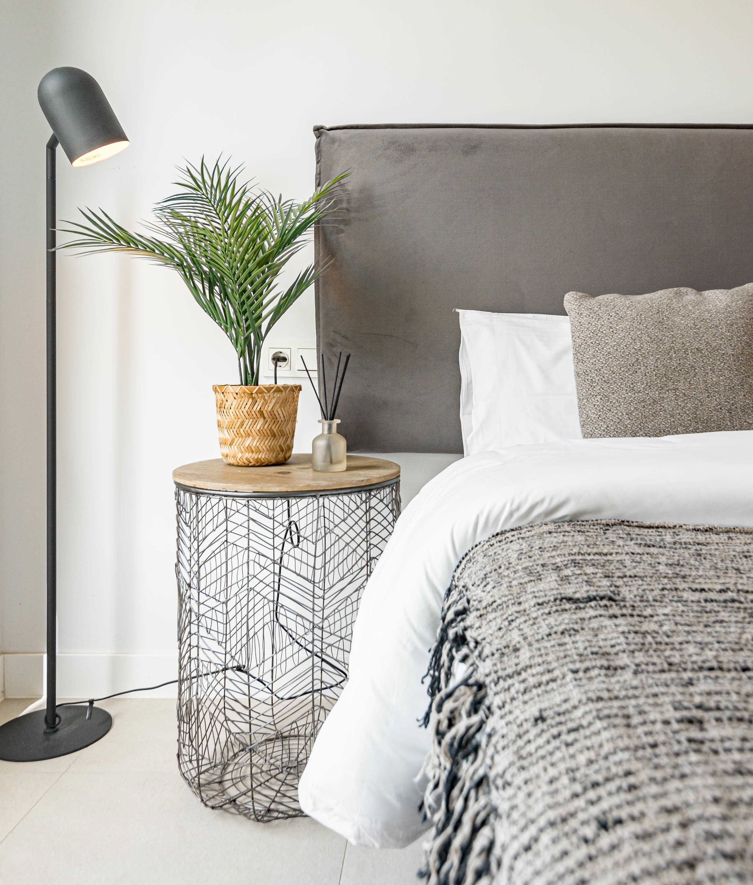 A neatly arranged bedroom with a bed, bedside lamp, potted plant, and a small round side table with a woven basket and reed diffuser.