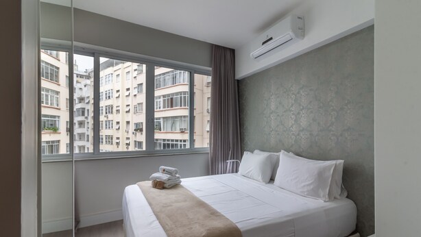 Une chambre d’hôtel avec une grande fenêtre, un lit avec du linge de lit blanc, un plaid beige et une table de chevet blanche.