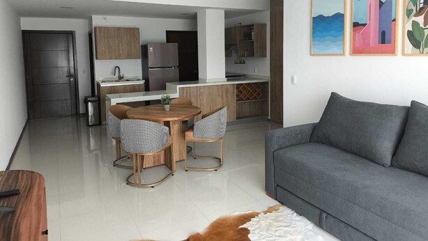 Ein modernes Wohnzimmer mit einem grauen Sofa, einem Esstisch mit karierten Stühlen und einer Küchenzeile mit Holdekor und einem Kühlschrank.