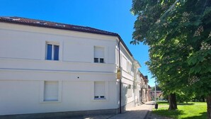 Un bâtiment blanc avec un toit recouvert de tuiles, un trottoir pavé et une zone herbeuse avec des arbres.