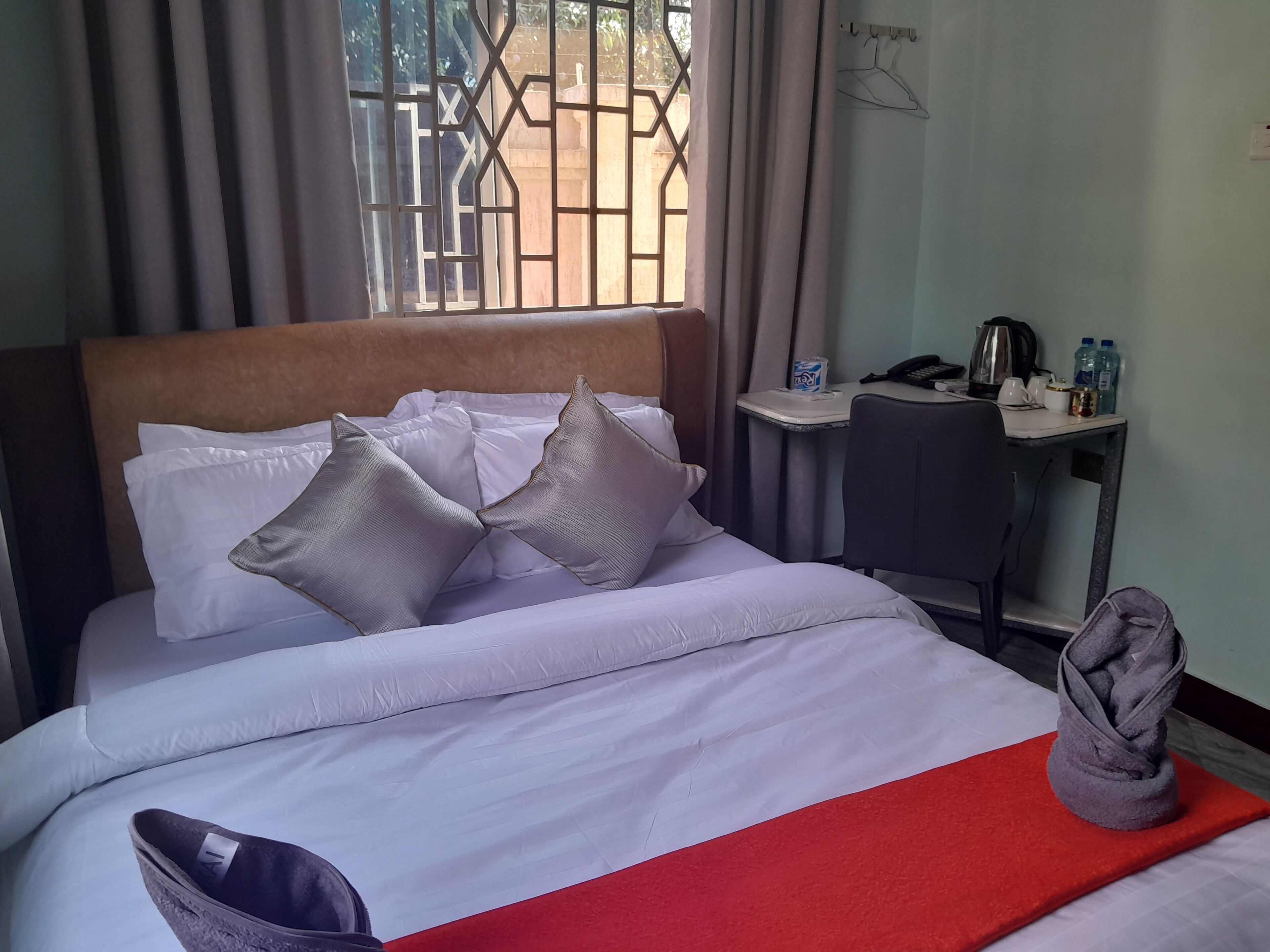 A neatly made bed with white linens and multiple pillows, a small desk with a chair, a kettle, and a water bottle.