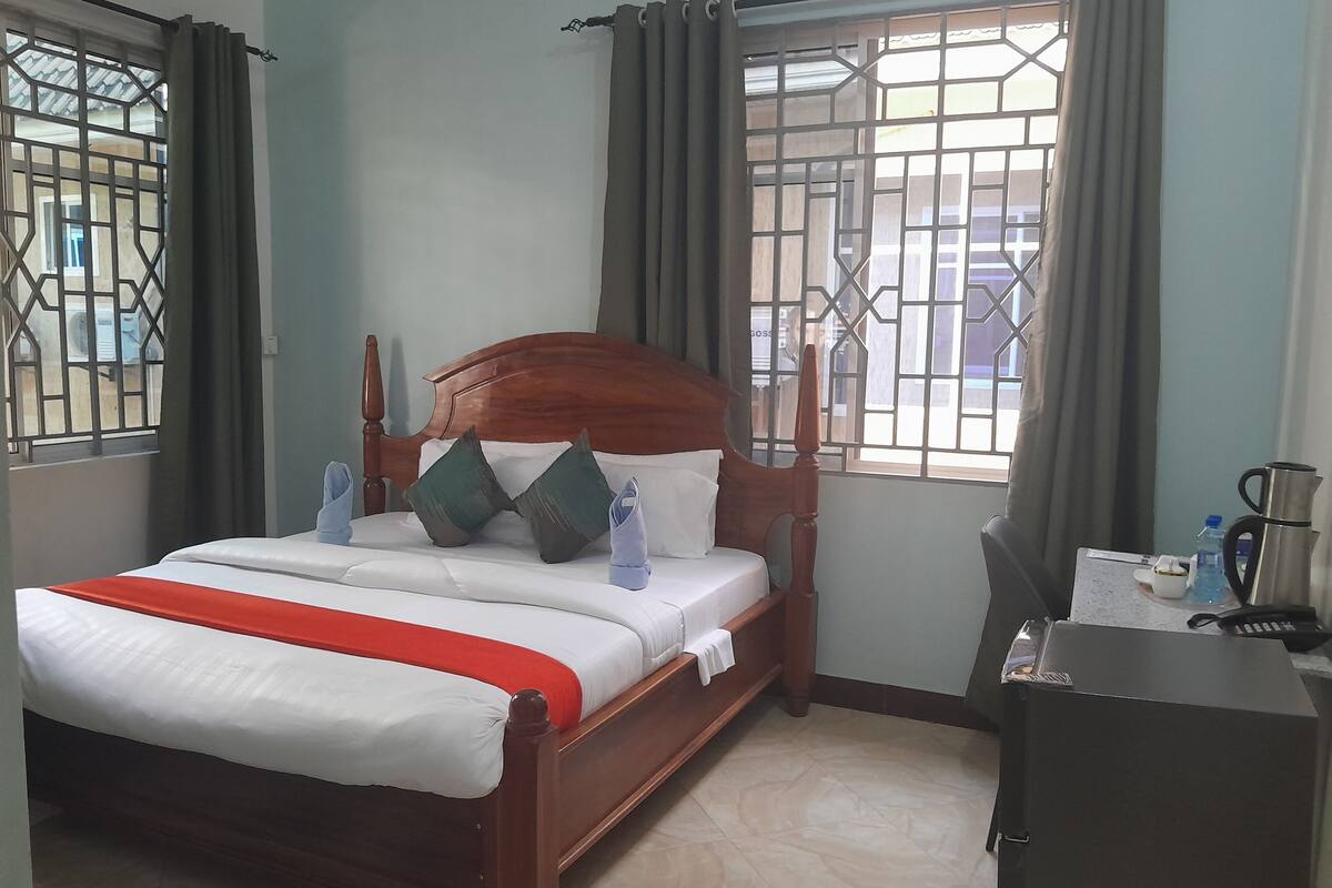 Una cama bien hecha con cabecera de madera, una mesita de noche con hervidor y una ventana con barrotes y cortinas.