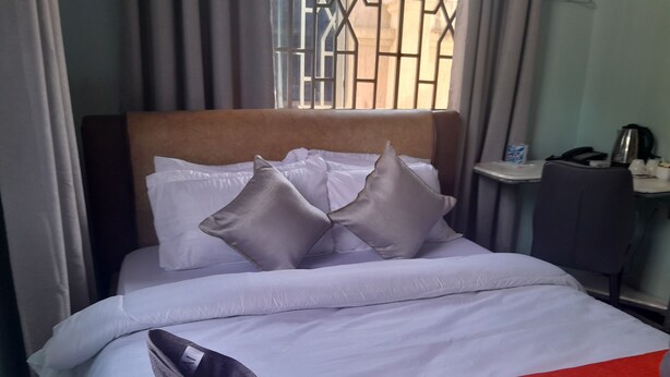 A neatly made bed with white bedding and two grey pillows, a red throw blanket, and a grey boot on the floor. - DAG HOTEL MOROGORO (Morogoro)