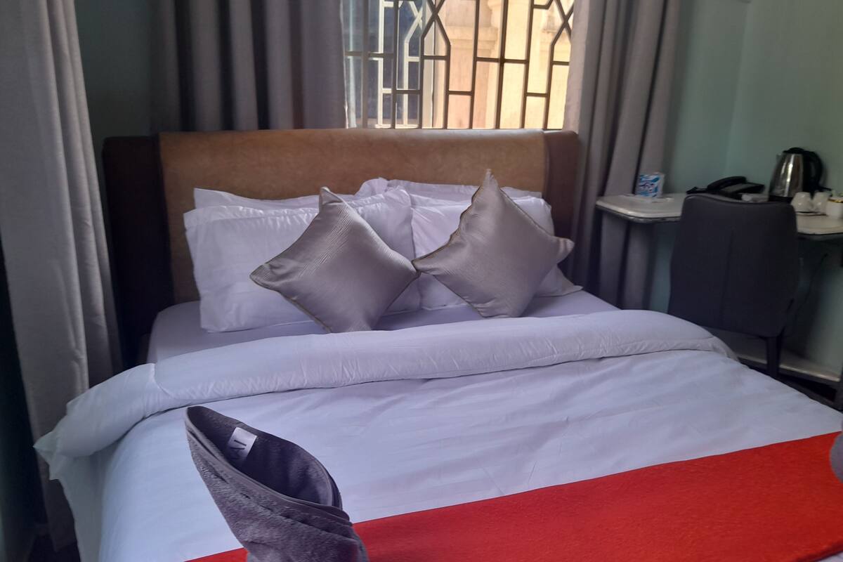 Una cama bien hecha con ropa blanca y dos almohadas grises, una manta roja y una bota gris en el suelo.