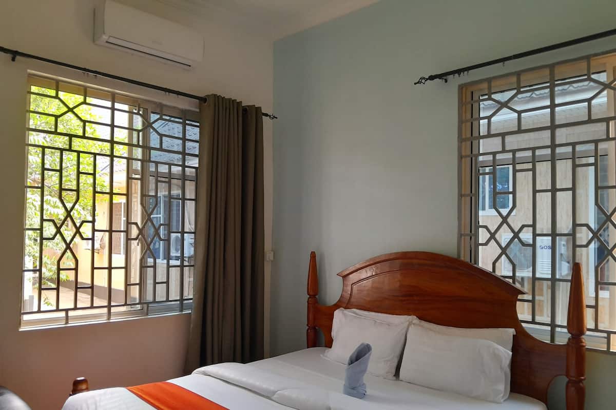 Una cama bien hecha con sábanas blancas y un plaid naranja, un cabecero de madera, y una ventana con celosía metálica y cortinas.