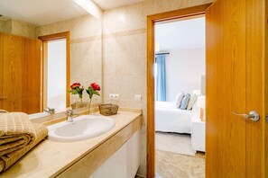 Une salle de bain avec un lavabo, un miroir et une porte en bois donnant sur une chambre à coucher équipée d’un lit et de rideaux.