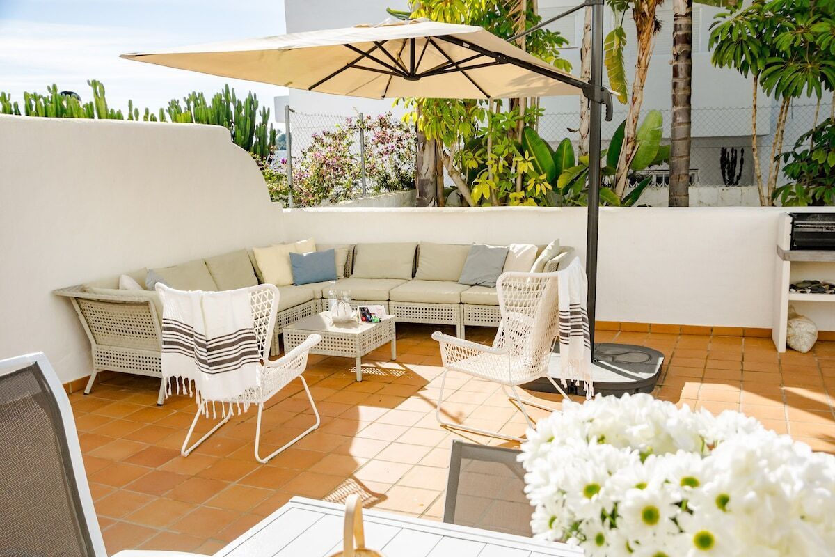 A white outdoor seating area with a sofa, chairs, and a table, surrounded by plants and a large umbrella.