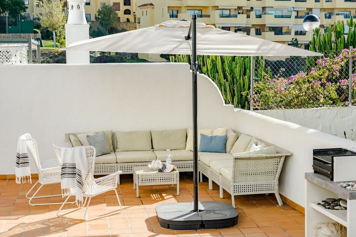 A rooftop terrace with a white sofa, wicker chairs, a round table, and a large umbrella.
