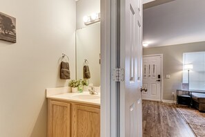 Un baño con tocador de madera, un espejo y un lavabo. Hay una puerta que conduce a otra habitación.