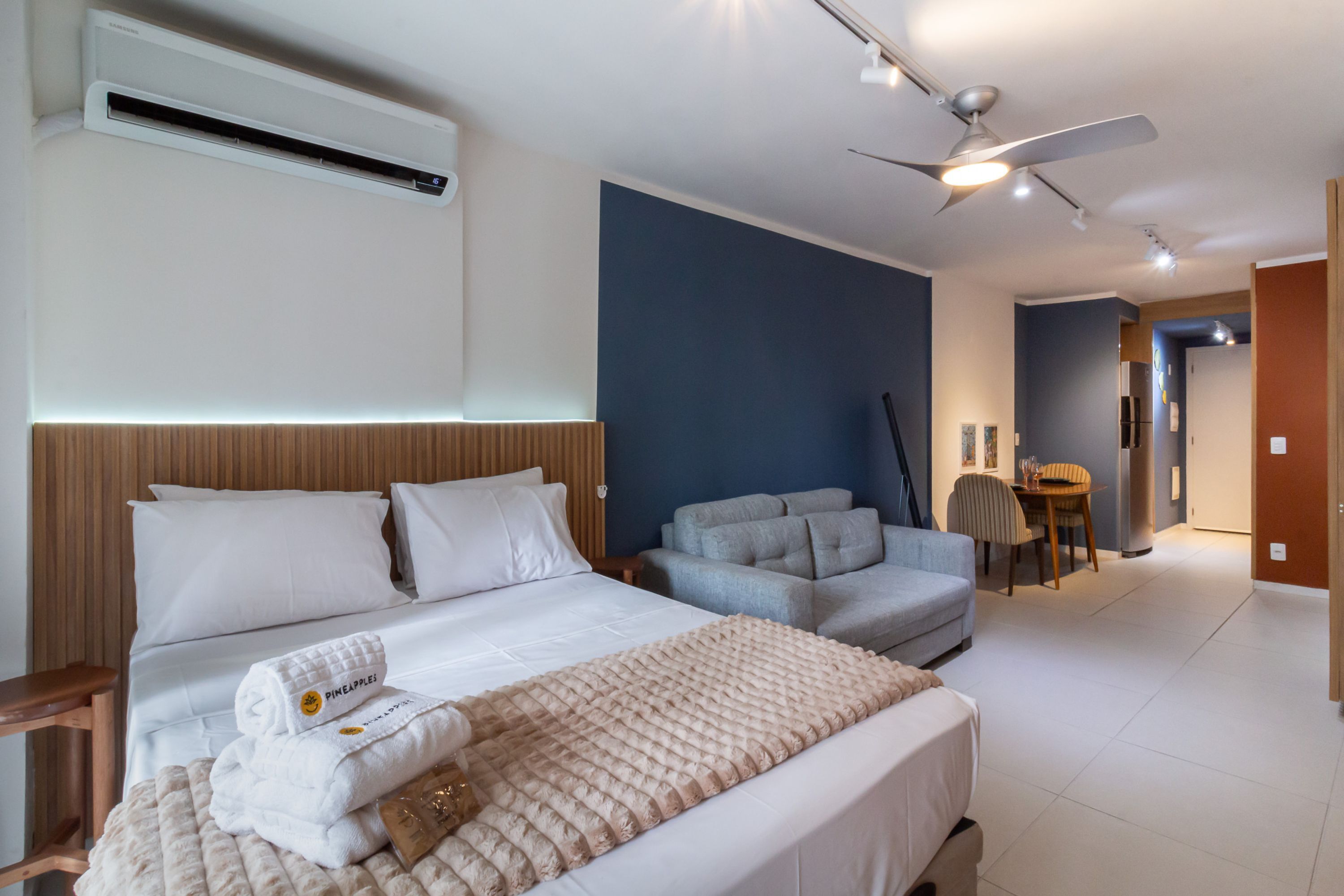A modern hotel room with a bed, a sofa, a dining table, and a ceiling fan.