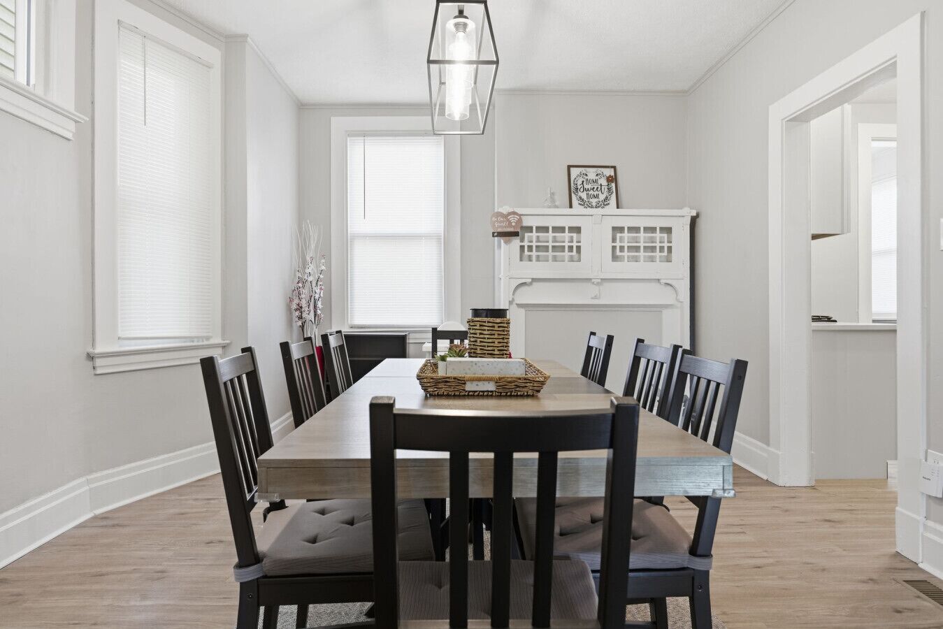A dining area with a long table, black chairs, and a chandelier. There is a white cabinet with glass doors in the background.