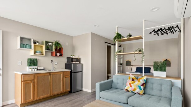A modern kitchen with wooden cabinets, a built-in microwave, and a refrigerator. There is a gray sofa with a colorful pillow and a wooden coffee table in the living area. - Quaint, New Hideaway Studio-sized Room (San Diego)
