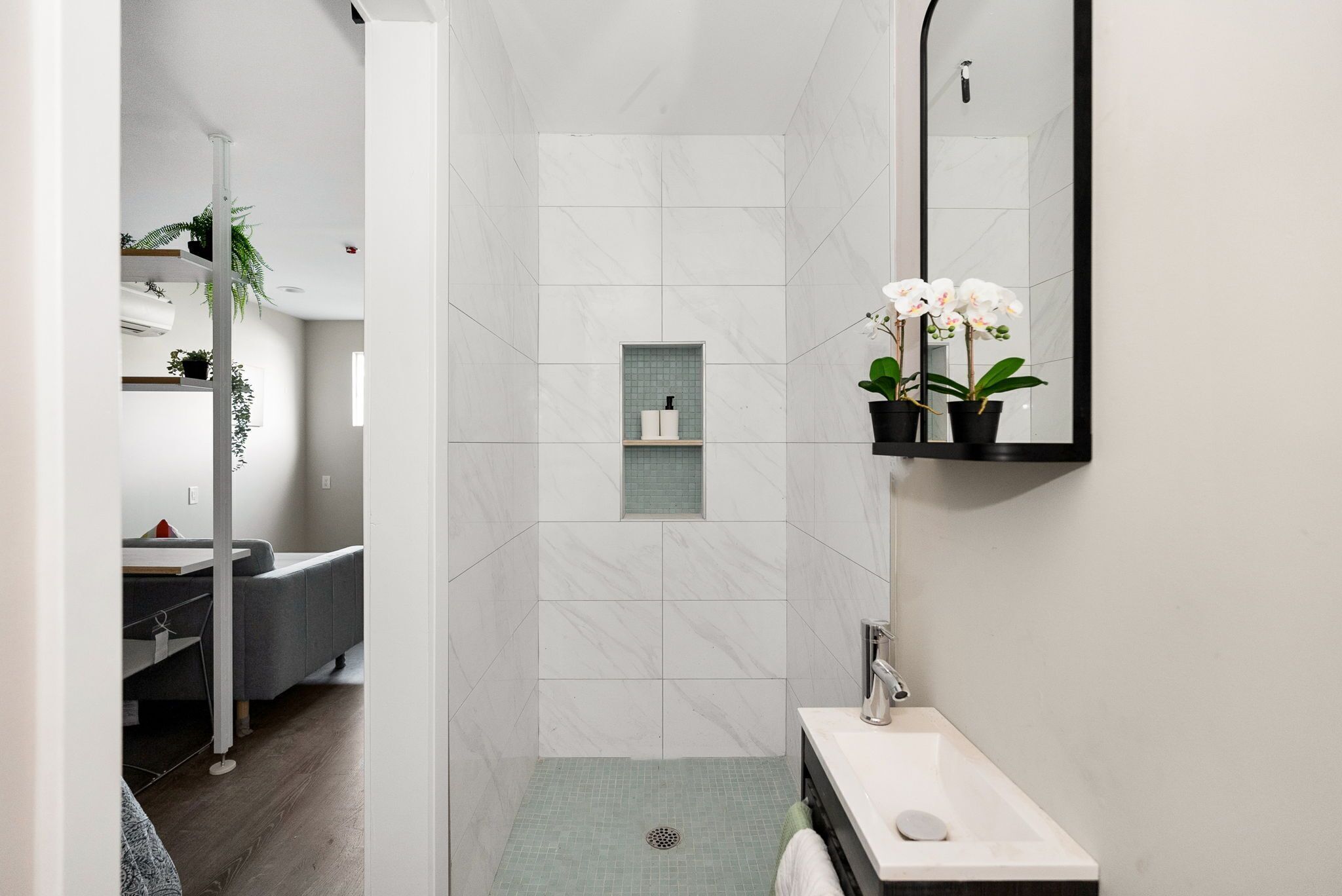 A modern bathroom with a shower, sink, and mirror. There are plants and a towel rack.