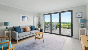 Un salon moderne avec un canapé gris, une petite table d’appoint en bois, un tapis bleu et une grande porte-fenêtre coulissante donnant sur une terrasse extérieure.