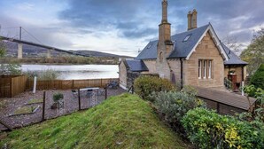 Une maison en pierre avec une grande cheminée, une terrasse en bois et une vue sur un pont enjambant une étendue d’eau.