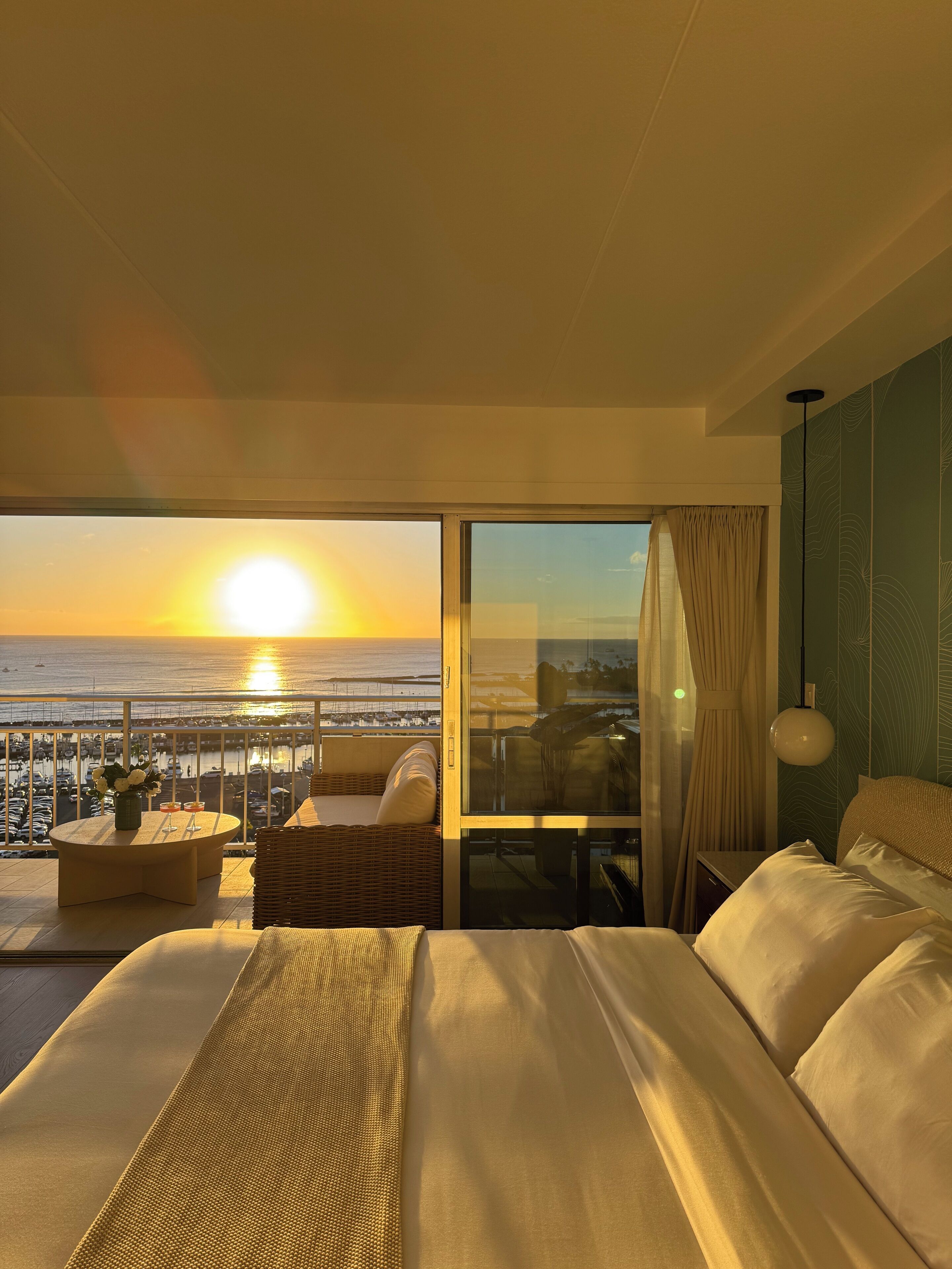 A hotel room with a large bed, a balcony with a view of the sunset over the water, and a dining area with a table and chairs.