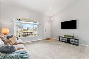 A living room with a large window, a flat-screen TV mounted on the wall, a sofa with cushions, and a small side table with a plant. - Cozy 3BR Nampa Home • Spacious Yard + Smart TV (Nampa)