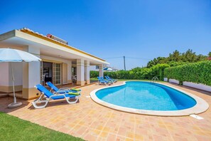 Ein Schwimmbecken mit Liegestühlen und Sonnenschirmen, eine überdachte Terrasse und ein gepflegter Garten.