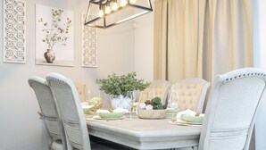 Un comedor con una mesa redonda, cuatro sillas tapizadas, un jarrón con flores, un frutero y un adorno de pared.