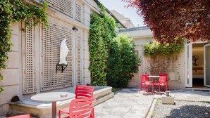 Un patio con sillas rojas y una mesa, rodeado por un edificio con celosía y follaje.