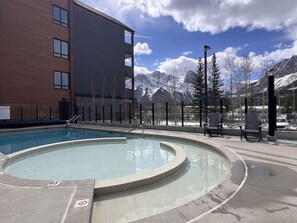 A swimming pool with a tiled edge, a building with a brick facade, and a mountainous backdrop. - NEW Condo w/ VIEWS — Sleeps 9 w/ Pool, Hot Tub! (Canmore)