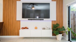 A modern living room with a flat-screen TV mounted on a white wall, a ceiling fan, a wooden accent wall, a white console table with decorative items, and a potted plant. - Cozy Princeton Getaway (Homestead)