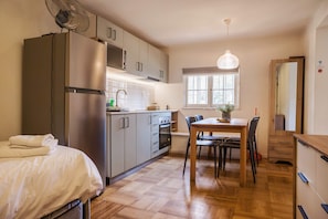 Cocina compacta con refrigerador, microondas, horno y lavaplatos. Hay una mesa de comedor con sillas, un ventilador y una ventana con luz natural.