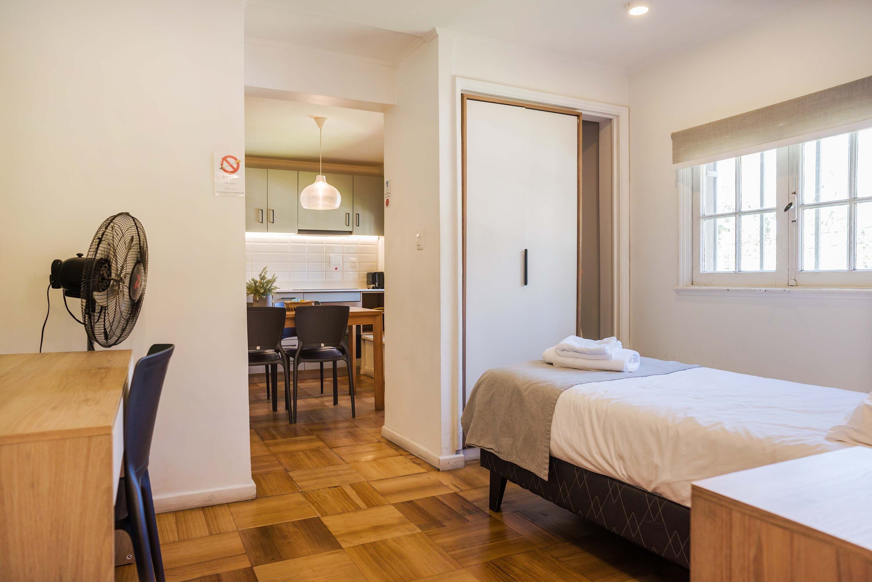 A compact hotel room with a bed, a desk, a chair, and a kitchenette in the background.