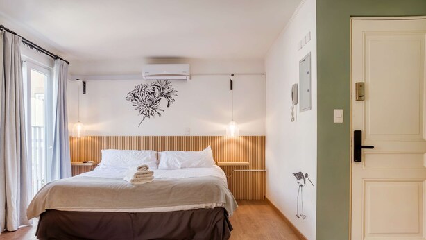 Ein ordentlich bezogenes Bett mit Holz-Kopfteil, eine wandmontierte Klimaanlage, eine Nachttischlampe und eine Tür mit Griff.
