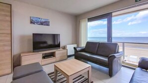 Habitación de hotel moderna con un ventanal grande que ofrece vistas al mar, un televisor de pantalla plana, un sofá de cuero y una pequeña mesa de madera.