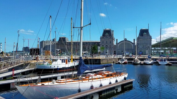 Ein Yachthafen mit vertäuten Booten, ein großes Gebäude im Hintergrund und ein klarer, blauer Himmel.