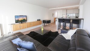 Ein modernes Wohnzimmer mit einer schwarzen Ledersofa, einem Flachbildfernseher, Holzschränken, einer Essecke mit Barhocker und einer Küche im Hintergrund.