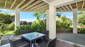Un patio techado con una mesa de vidrio y sillas de mimbre, rodeado de abundante vegetación y un cielo despejado.