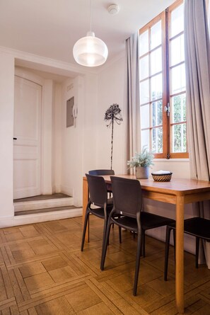A dining area with a wooden table, two chairs, a hanging light fixture, a window with curtains, and a door. - Chic Studio in Salvador Sleeps 4 Near Metro (Providencia)