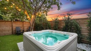 A hot tub in a backyard with a wooden fence, a tree, and string lights.