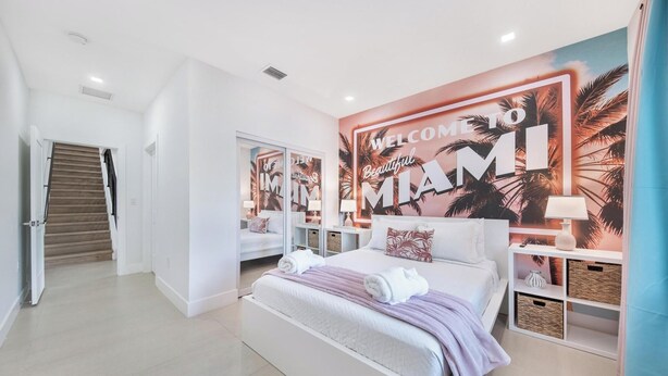 Una habitación de hotel moderna con una cama grande, una mesita de noche, una lámpara y un mural de temática tropical en la pared.