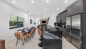 Una cocina moderna con un refrigerador de acero inoxidable grande, un comedor con una mesa larga y sillas, y vistas al exterior a través de amplios ventanales.